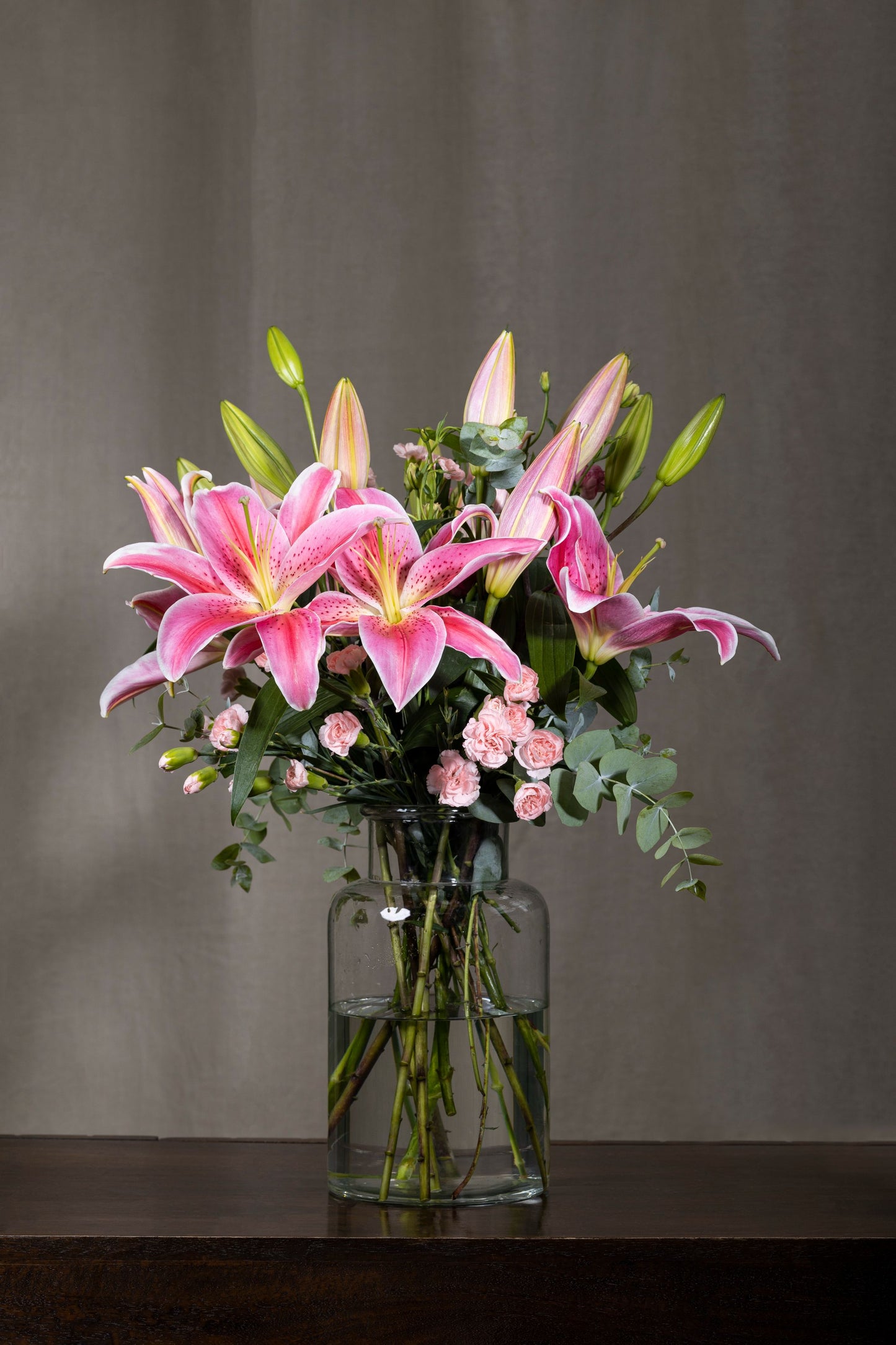 Pink Charming - Elegant glass biscuit jar arrangement with pink Oriental Lilies, aromatic silver dollar eucalyptus sprigs, and Spray Carnation flowers | Flower delivery in Bengaluru | Buy flower bouquets online Bengaluru | Same-day flower delivery Bengaluru | Online florist Bengaluru | Order flowers online Bengaluru | Luxury flower bouquet in Bengaluru