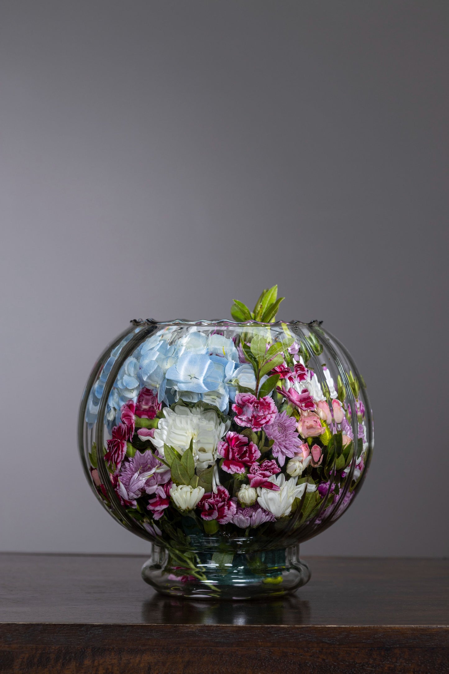 Elegant glass fishbowl with spray carnations and blue hydrangeas – a vibrant floral arrangement full of life and beauty