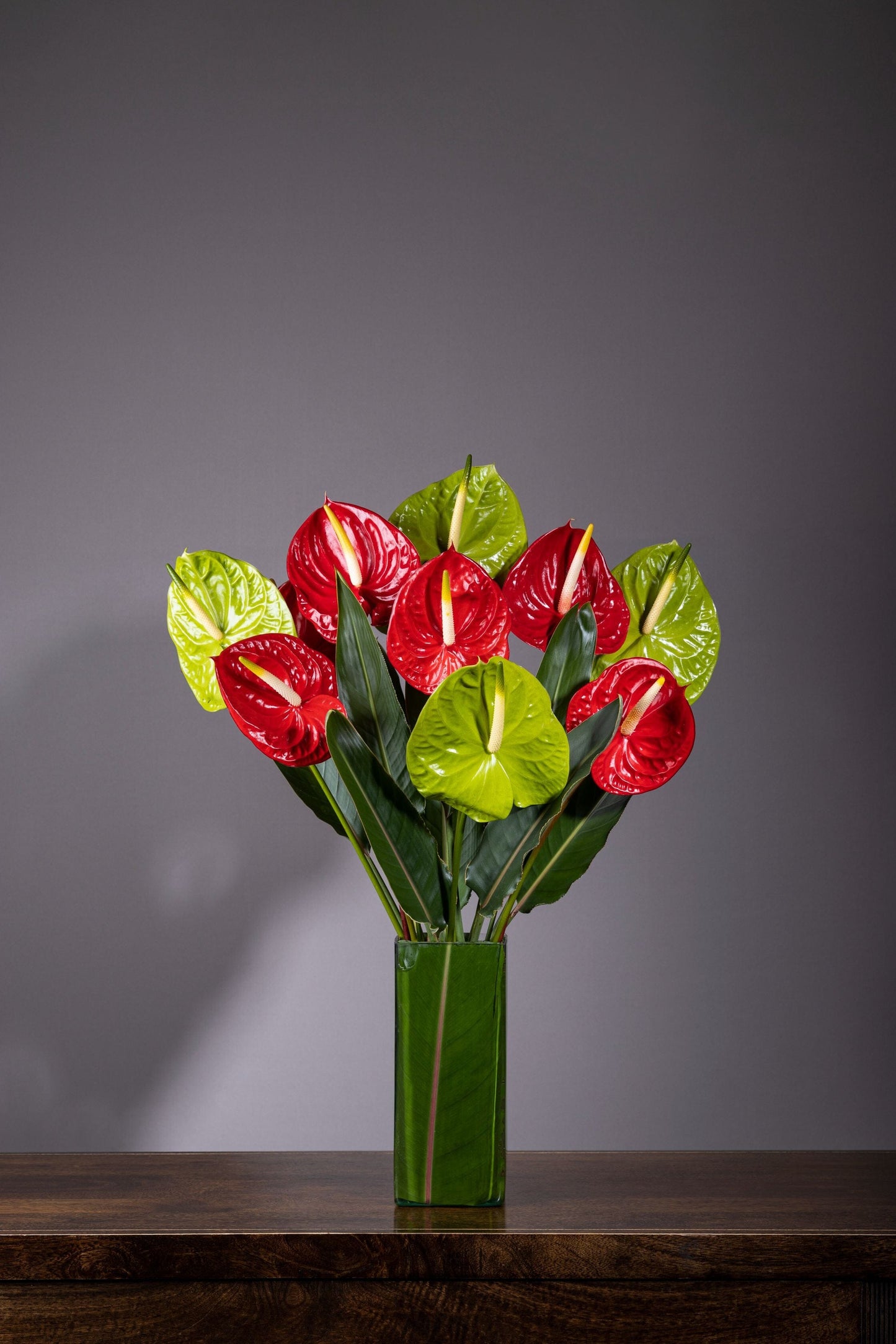Enchanting Hearts – A tall glass vase filled with vibrant red and green Anthurium flowers, featuring their signature heart-shaped blooms, complemented by lush foliage for a stunning floral display