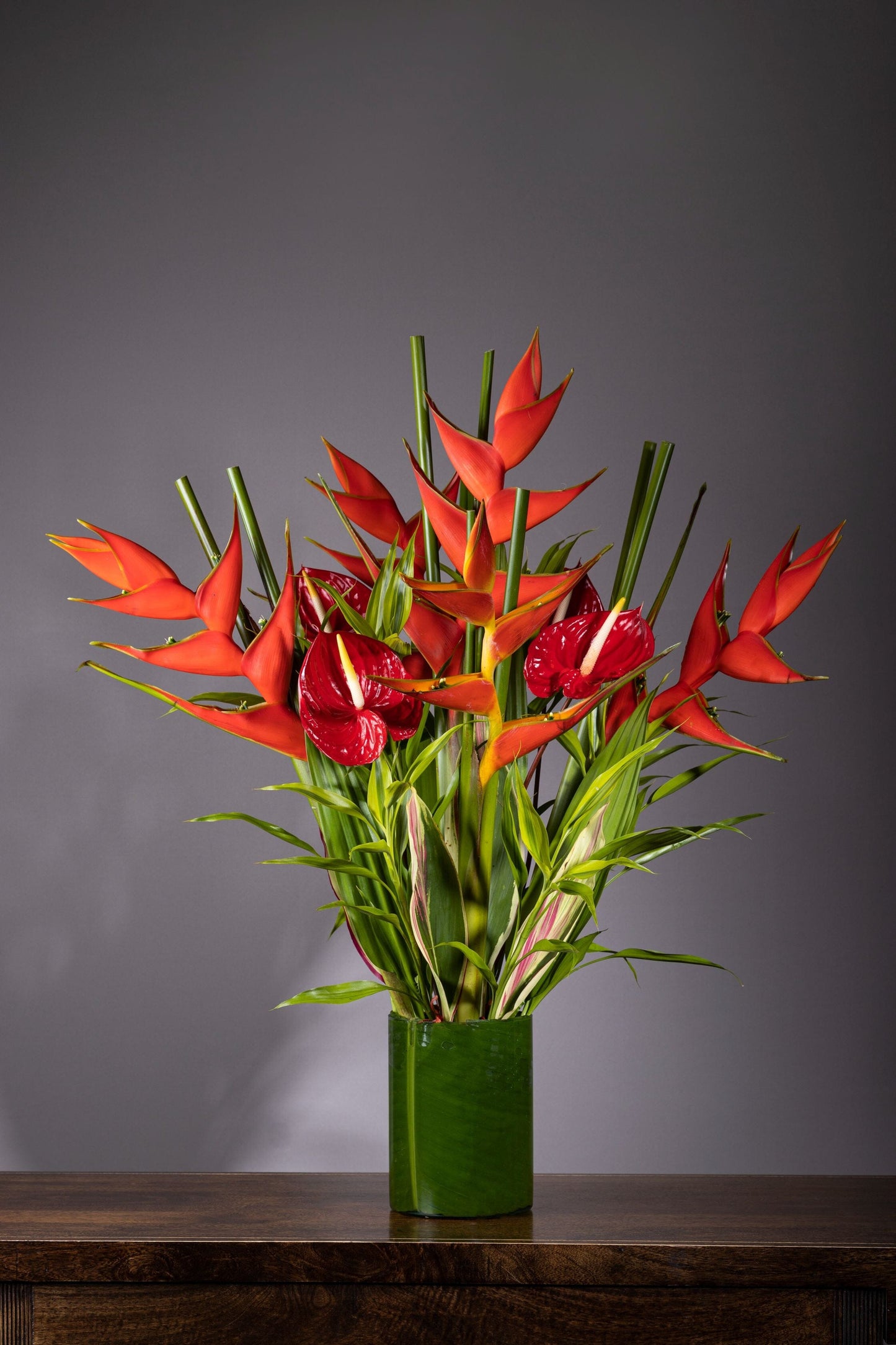 Rhapsody flower in bengaluru – A medium-height glass vase lined with lush leaves, showcasing tall and majestic Heliconia flowers in a striking fiery red hue, accented with unique foliage